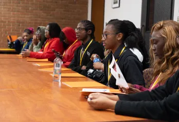 Students in Legislative Session – A diverse group of high school students dressed in business attire sits in a large lecture hall, attentively listening and taking notes as a student stands at a podium presenting an ordinance.