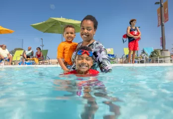 A mother and her toddler and child swim in a YMCA outdoor pool supervised by a YMCA Lifeguard