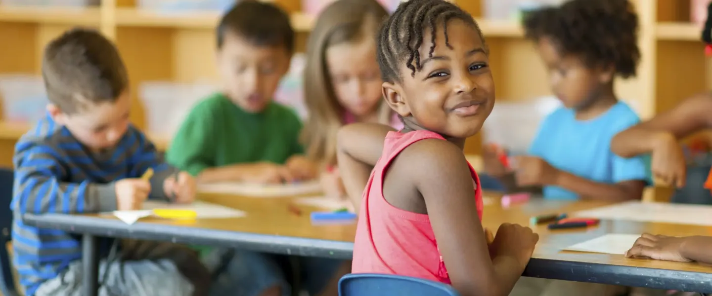 A young African American girl poses looking back towards the camera with a soft smile on her face. She is captured sitting, coloring pieces of papers with colorful markers with a diverse group of her classmates at a table in a classroom at the YMCA.