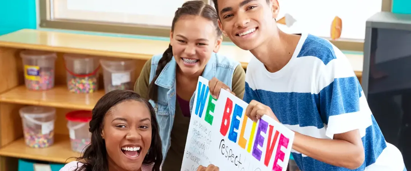An image of three teens making a colorful sign together that says, "we believe," as well as other words like "respect." The African American female, Asian female, and Middle Eastern male are all looking at the camera, smiling.