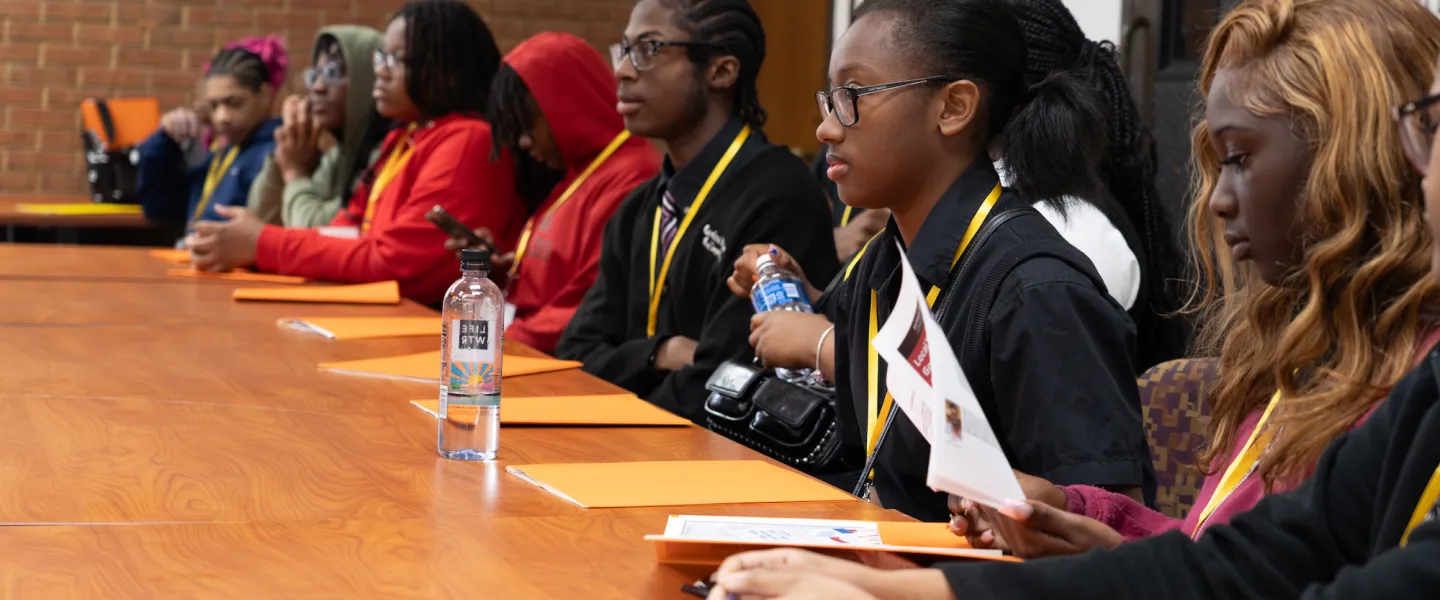 Students in Legislative Session – A diverse group of high school students dressed in business attire sits in a large lecture hall, attentively listening and taking notes as a student stands at a podium presenting an ordinance.