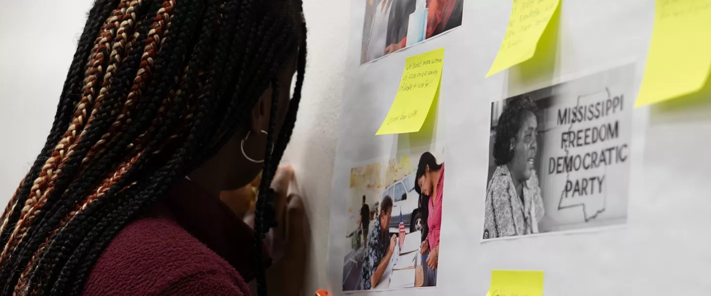 A female student writes on a yellow sticky note and places it on a whiteboard covered with other colorful notes. She is focused, with a pen in hand, contributing to a brainstorming or discussion activity.