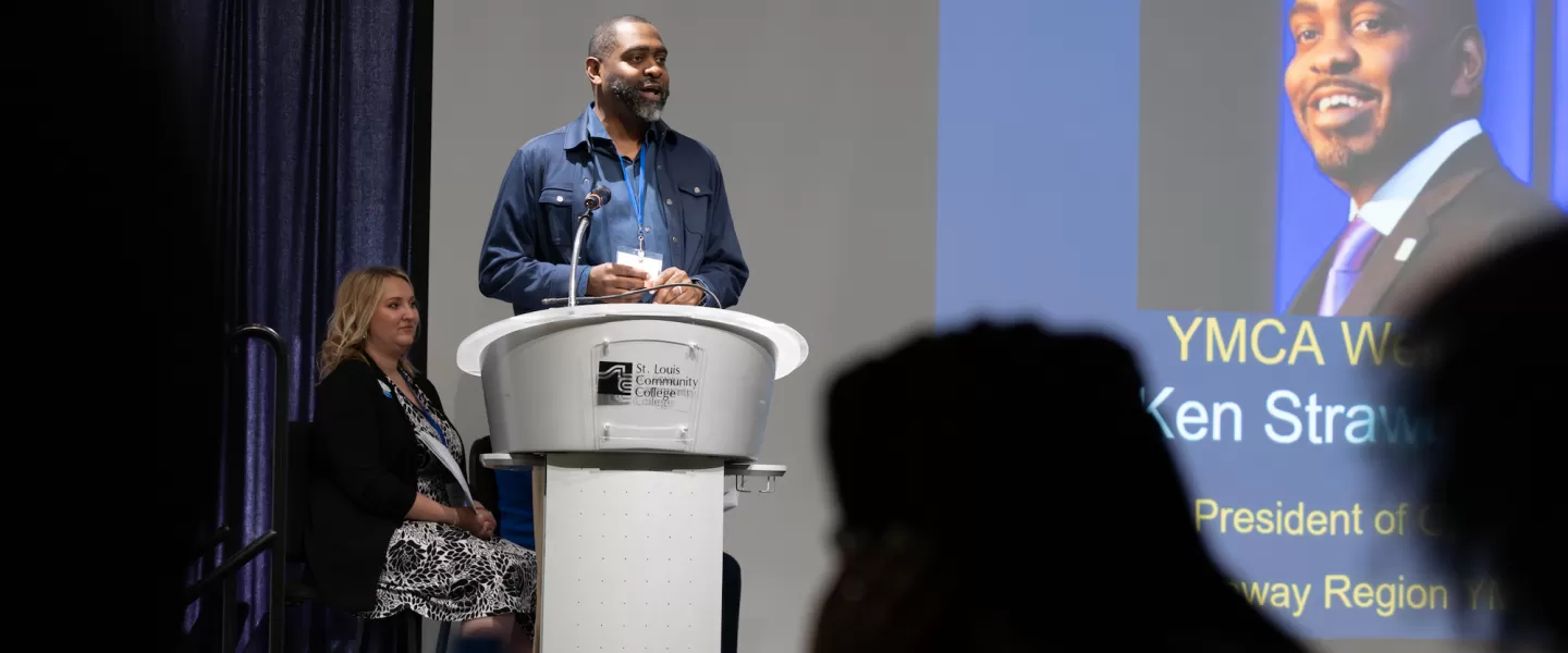 Speaker Addressing Students – A guest speaker, Ken Strawbridge, District Vice President at the Gateway Region YMCA, stands at the front of the room at a St. Louis Community College podium, delivering remarks to an audience of engaged students.