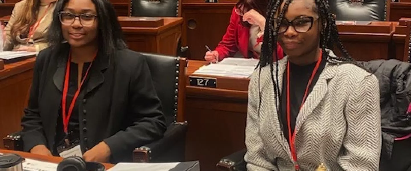 Two female students sit at desks on a courtroom floor, smiling at the camera. They are dressed in business attire, with papers and notebooks in front of them, participating in the Youth and Government conference.