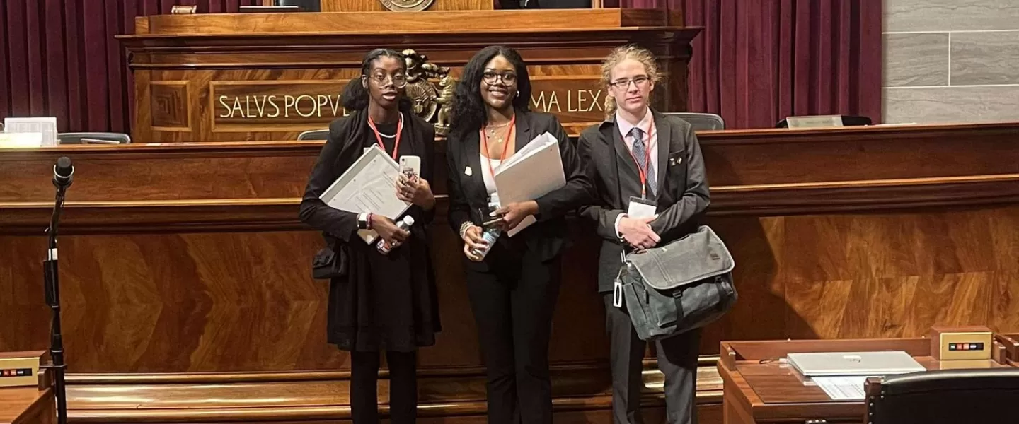 Students in Legislative Session – A diverse group of high school students dressed in business attire standing in front of the courtroom judge's podium.