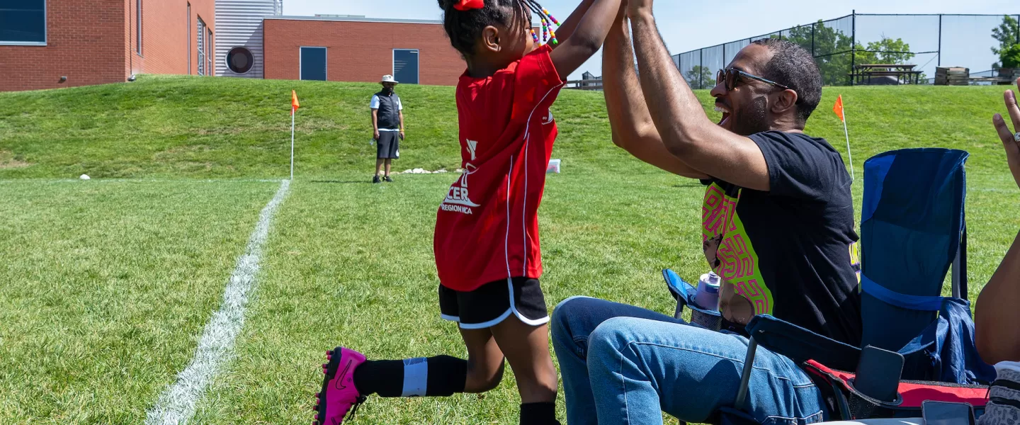 A young girl at the CITY SC Kickoff event high-fives her father on the sidelines as he cheers her on during the youth soccer game.