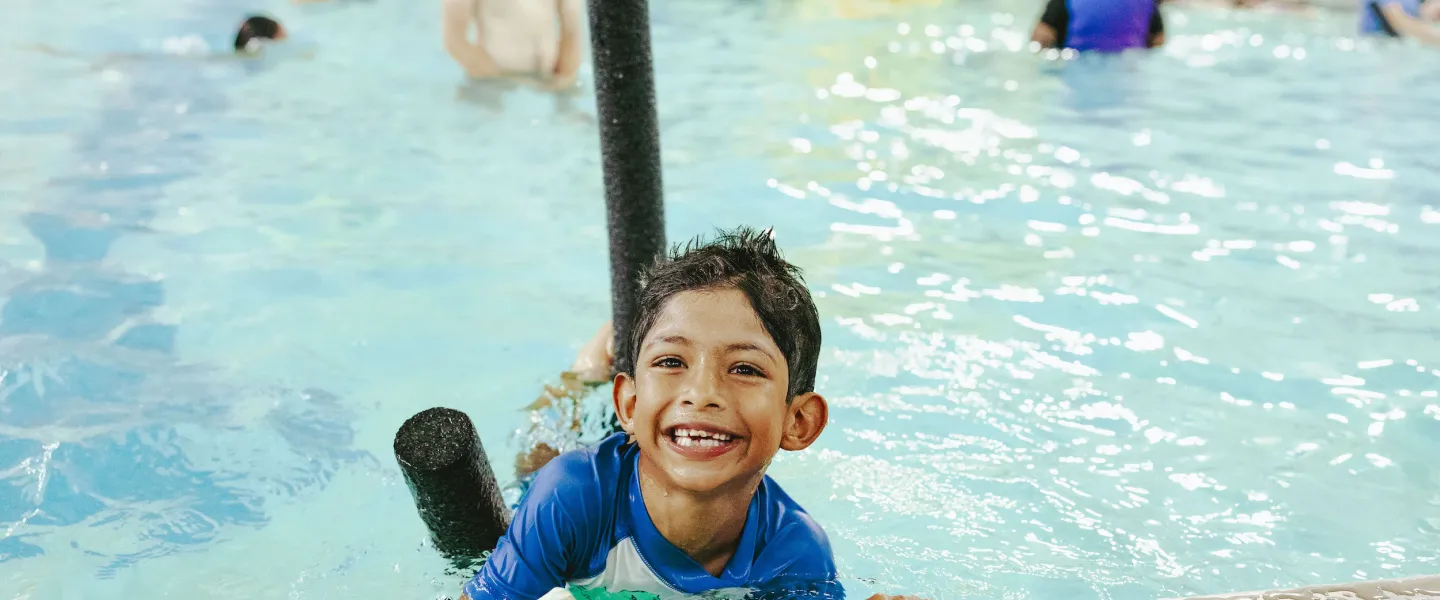 A young boy smiles at the edge of a YMCA indoor pool with a pool noodle showing off his green wrist band indicating he passed the YMCA's Go for Green Water Safety Program.