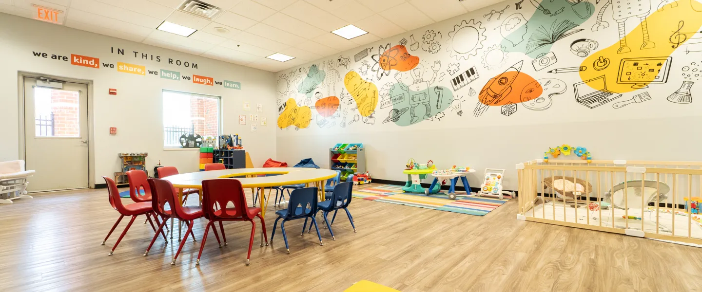 Modern YMCA Child Watch room featuring a large, colorful mural with science and space themes, a wooden table with red and blue chairs, a gated infant area, and a rainbow-striped rug.
