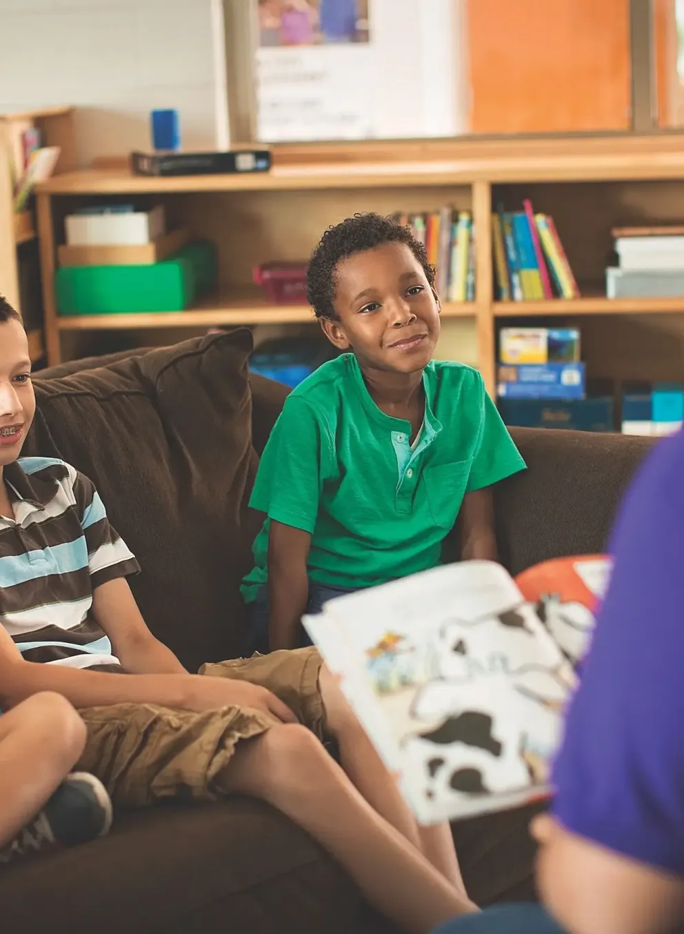 A female YMCA staff member wearing a purple shirt reads a book to three children, one Hispanic girl, one Caucasian boy, and an African American boy. All three children are happily listening to the staff member read to them while they sit on a couch in their classroom.