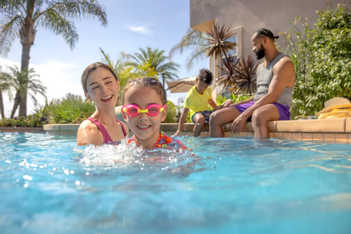 A family swims in an outdoor pool with a child wearing bright pink goggles and mother smiling next to her