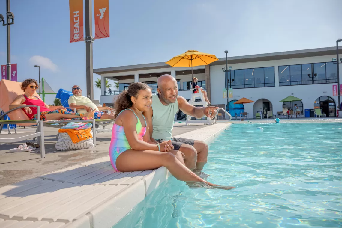 A father teaching his daughter about the "Reach, Throw, Don't Go" water safety technique by the edge of a YMCA outdoor pool