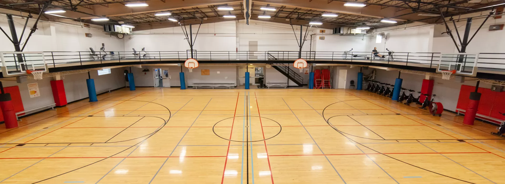 Gymnasium with multiple hoops. Small bench seats along walls of court. Large indoor track running overhead with machines in corners.