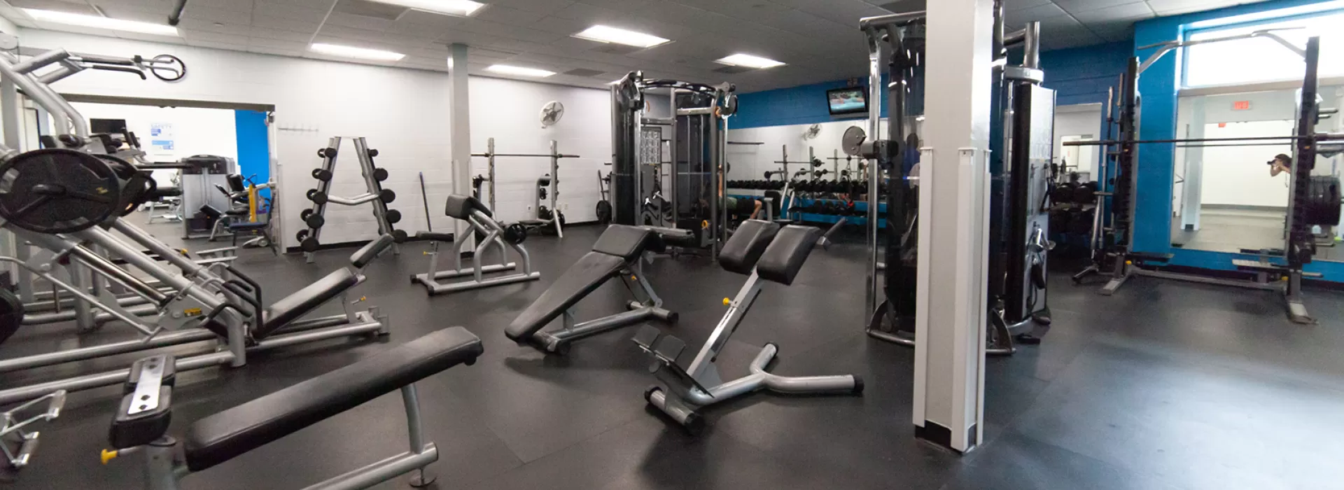 Weight room showing squat racks, free weights, and benches. Industrial feeling room with dark flooring and white and blue walls.