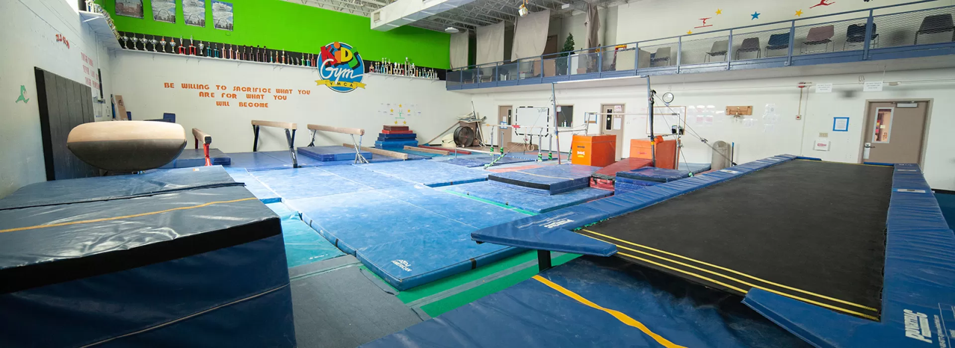Gymnastics room with balance beams, trampoline, parallel bars, and soft floors. Upstairs viewing area for parents.