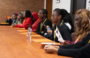 Students in Legislative Session – A diverse group of high school students dressed in business attire sits in a large lecture hall, attentively listening and taking notes as a student stands at a podium presenting an ordinance.