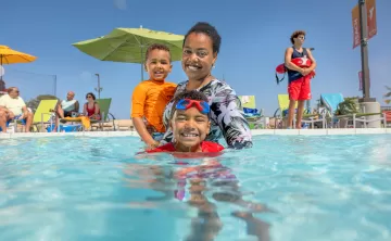 A mother and her toddler and child swim in a YMCA outdoor pool supervised by a YMCA Lifeguard